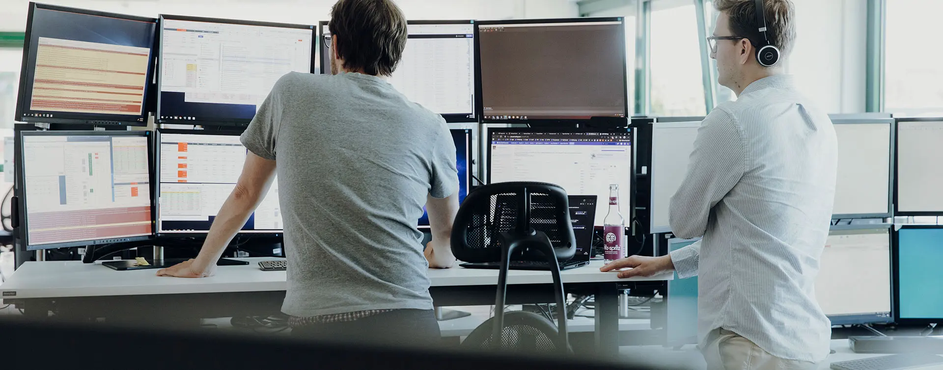 Zwei Trader stehen an einem höhenverstellbaren Schreibtisch, auf dem 8 Monitore montiert sind und überwachen den Handel
